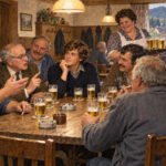 Sieben Menschen sitzen in einer gemütlichen Kneipe um einen Holztisch herum, lachen und unterhalten sich bei einem Bier, während der warme Schein der Kameradschaft den Raum erfüllt. Neben ihnen steht eine lächelnde Frau mit einem Tablett voller weiterer Getränke, während im Hintergrund leise lebhafte Stammtischmelodien erklingen. An der Wand hängen Mäntel, und durch ein Fenster blickt man auf ein malerisches Dorf draußen.