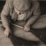 Eine ältere Person mit Mütze und Jacke sitzt an einem Holztisch und hält zwei handgefertigte Besen in der Hand. Das sepiafarbene Foto, das von oben aufgenommen wurde, erinnert an die Stille des Winters und hebt die Texturen des Holzes und der Besen hervor.