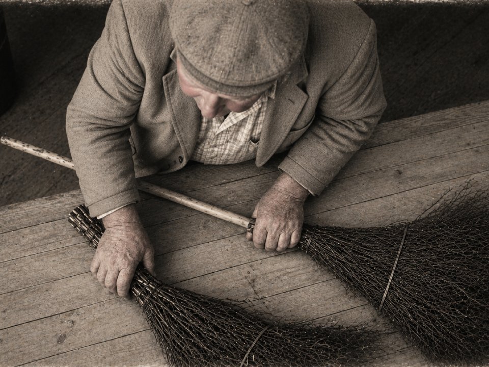 Eine ältere Person mit Mütze und Jacke sitzt an einem Holztisch und hält zwei handgefertigte Besen in der Hand. Das sepiafarbene Foto, das von oben aufgenommen wurde, erinnert an die Stille des Winters und hebt die Texturen des Holzes und der Besen hervor. Eine ältere Person mit Mütze und Jacke sitzt an einem Holztisch und hält zwei handgefertigte Besen in der Hand. Das sepiafarbene Foto, das von oben aufgenommen wurde, erinnert an die Stille des Winters und hebt die Texturen des Holzes und der Besen hervor.