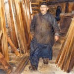 Ein älterer Mann in einer langen, dunklen Robe und einer Kappe geht durch eine Werkstatt, in der Holzbretter aufgestapelt sind, während im Hintergrund Regale und Werkzeuge zu sehen sind – vielleicht führt er damit das Handwerk seiner erstaunlichen Vorfahren fort.