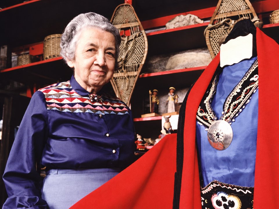 Gladys Tantaquidgeon steht neben einer Schaufensterpuppe, die ein traditionelles blaues Gewand mit einem roten Umhang und einer großen silbernen Brosche trägt; im Hintergrund stehen Holzschneeschuhe auf einem Regal. Gladys Tantaquidgeon steht neben einer Schaufensterpuppe, die ein traditionelles blaues Gewand mit einem roten Umhang und einer großen silbernen Brosche trägt; im Hintergrund stehen Holzschneeschuhe auf einem Regal.